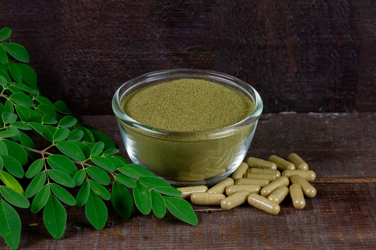 Moringa oleifera powder in transparent bowl with green leaf and capsule on rustic wooden table.