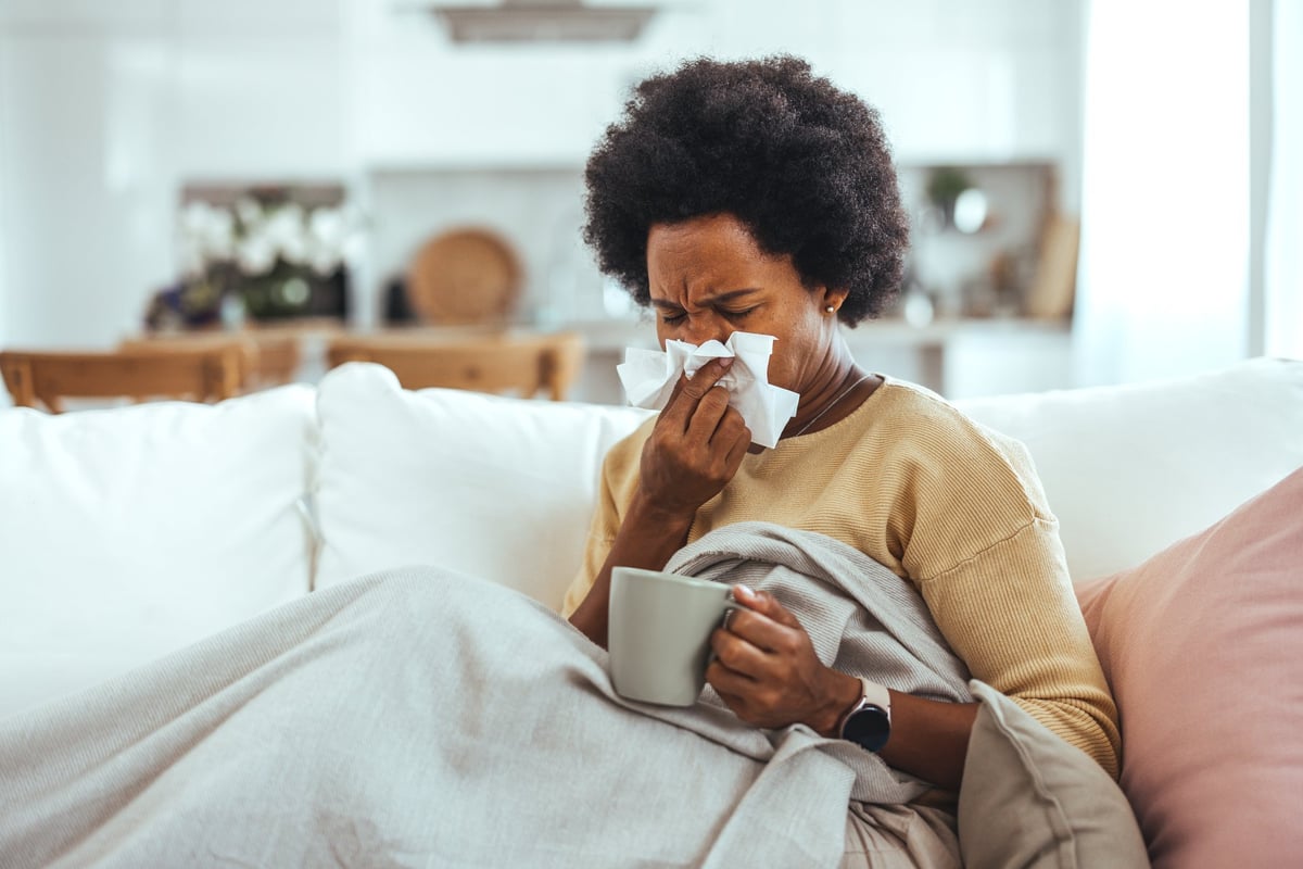 Sick young woman sitting on sofa blowing her nose at home.