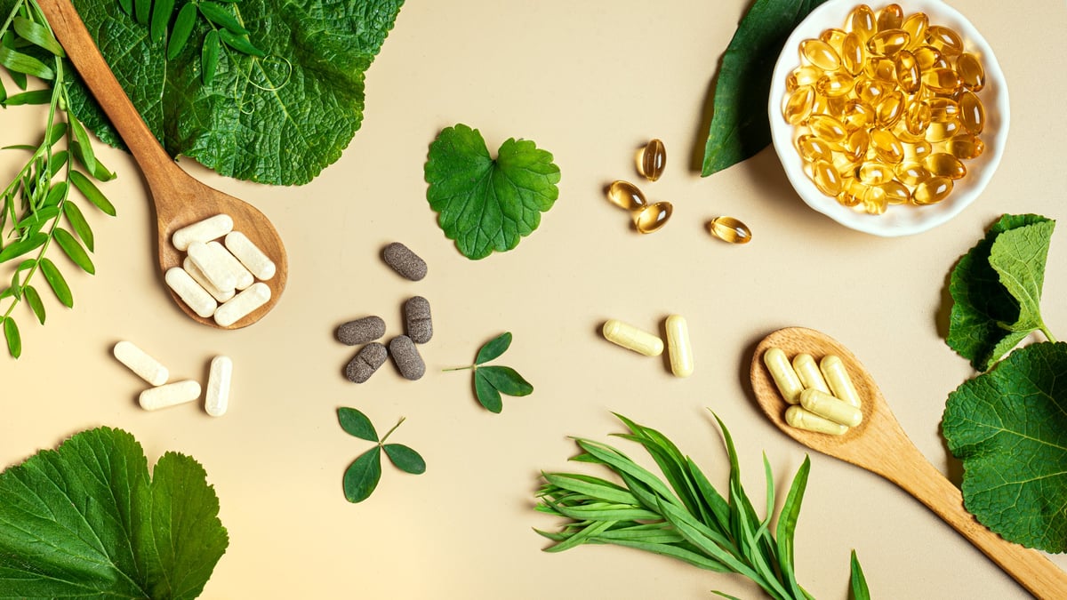 Vegan vitamin capsules for immunity support and healthy lifestyle on light beige background. Natural softgels and pills supplements and various green plant leaves, banner.