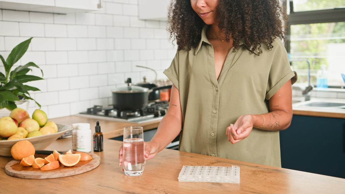 A woman stands at a kitchen counter holding a glass of water and a vitamin pill beside a pill organizer, fresh oranges and supplements.